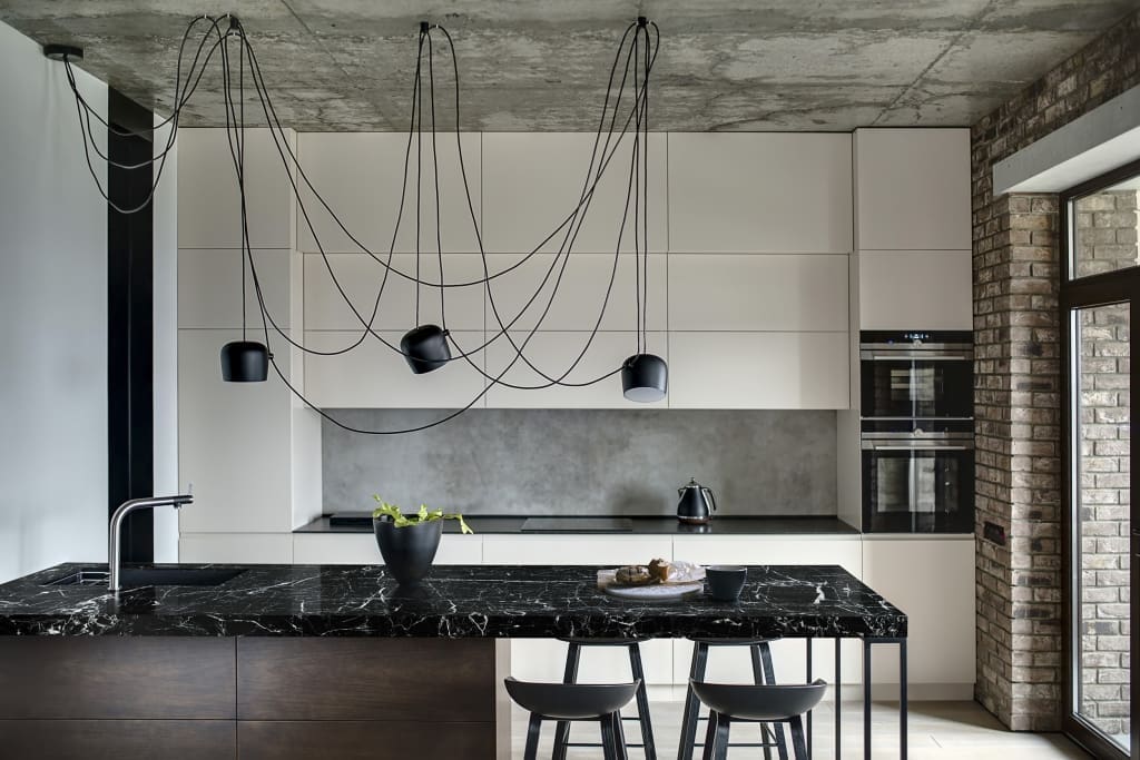 Kitchen in a loft style with concrete and brick walls. There is a kitchen island with a sink, plant and black chairs, light lockers with built-in oven and fridge, dark worktop with stove and teapot. Plan-It Stone, Best kitchen worktops in Bristol.